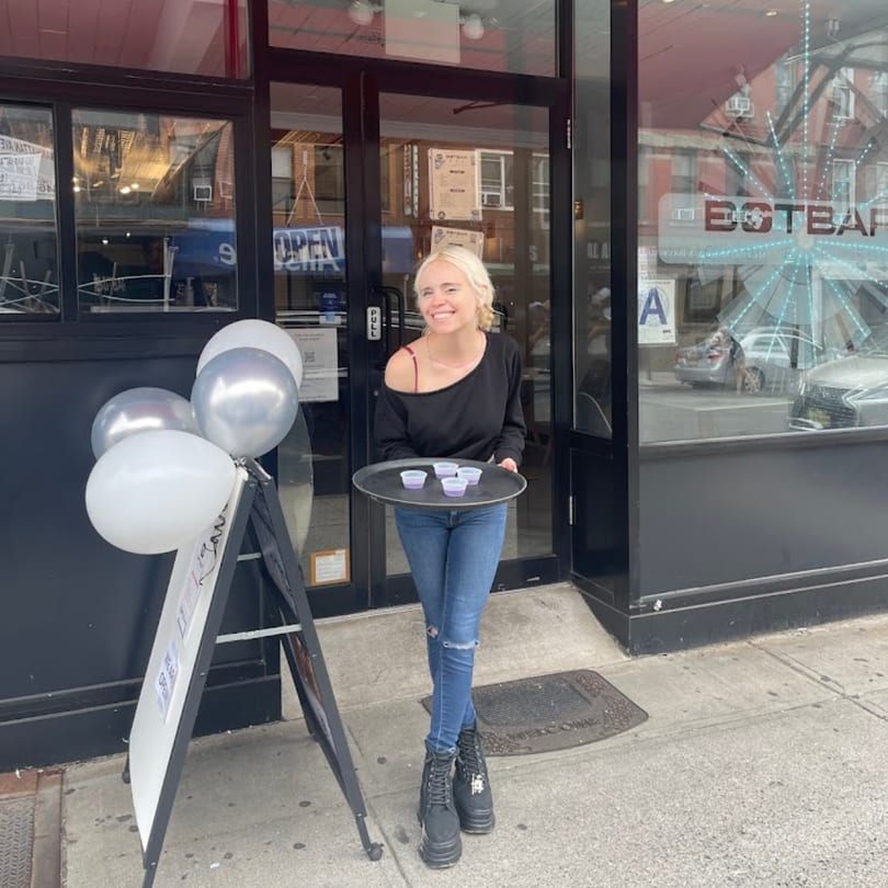 A smiling woman in jeans and a black top holds a tray of samples outside Botbar Vegan Cafe.