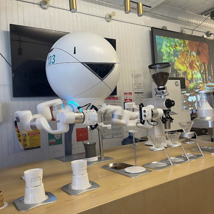 A white robotic barista with multiple arms preparing coffee on a light wooden counter.
